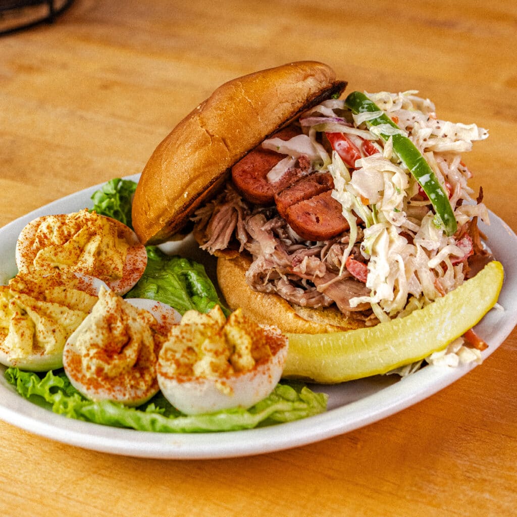 Succulent Oklahoma BBQ pulled pork sandwich topped with coleslaw and smoked sausage, served with a side of deviled eggs at Earl's Rib Palace. Enjoy a hearty, flavorful meal perfect for BBQ lovers.