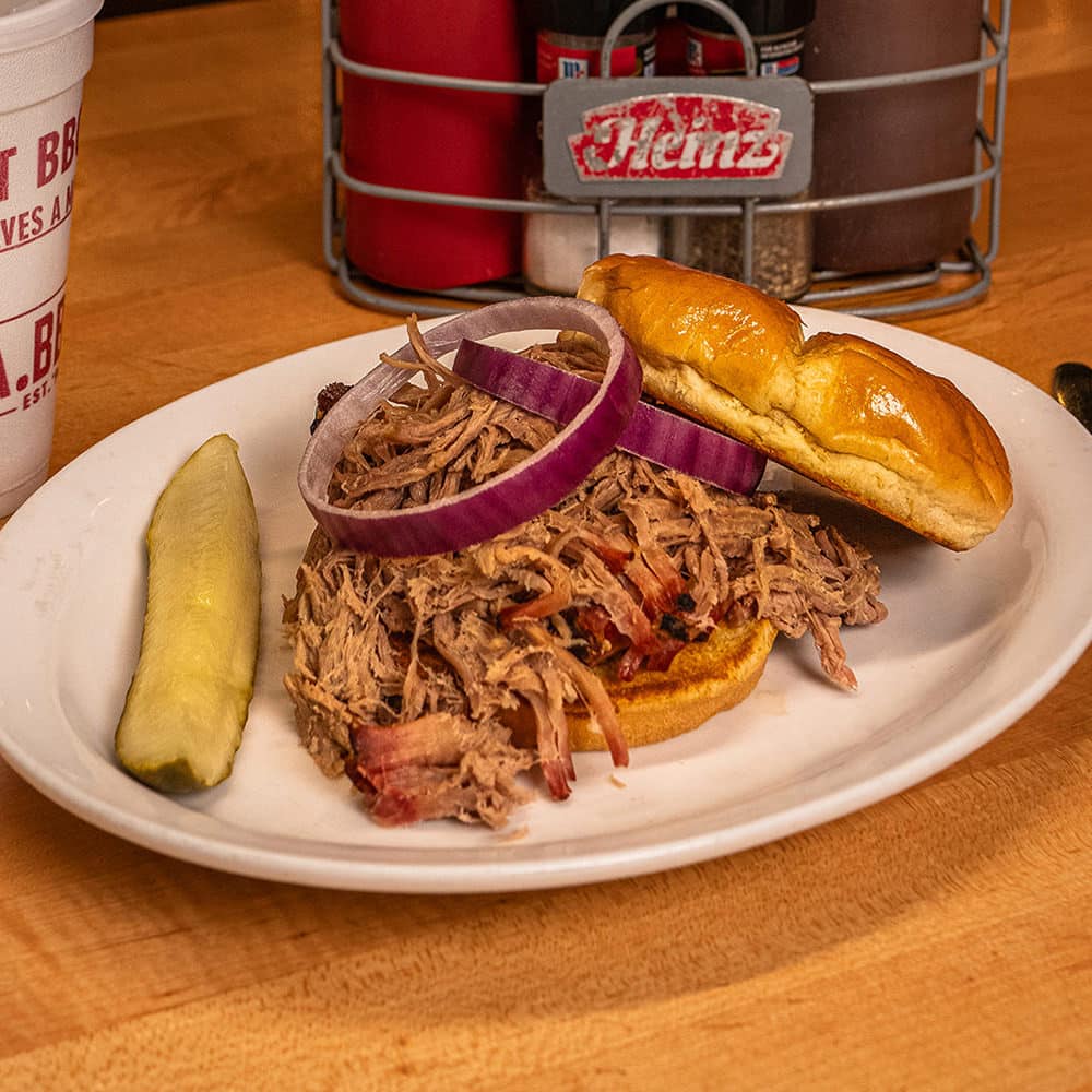 Pulled pork sandwich topped with red onions, served with a pickle spear, showcasing Earl's Rib Palace's signature Oklahoma BBQ flavors. Perfectly paired with a refreshing drink, this dish highlights the authentic, mouthwatering BBQ experience.